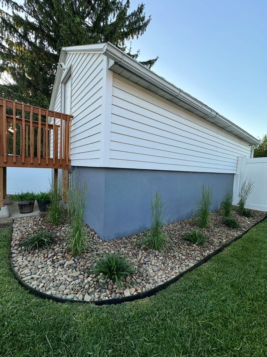 Side view of a house with white siding, landscaped with rocks and greenery, near a wooden deck.
