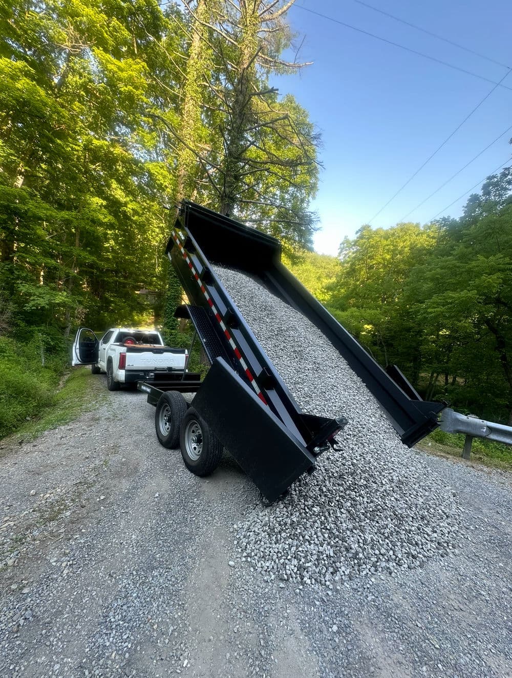 Dump truck unloading gravel on a gravel road surrounded by lush green trees.